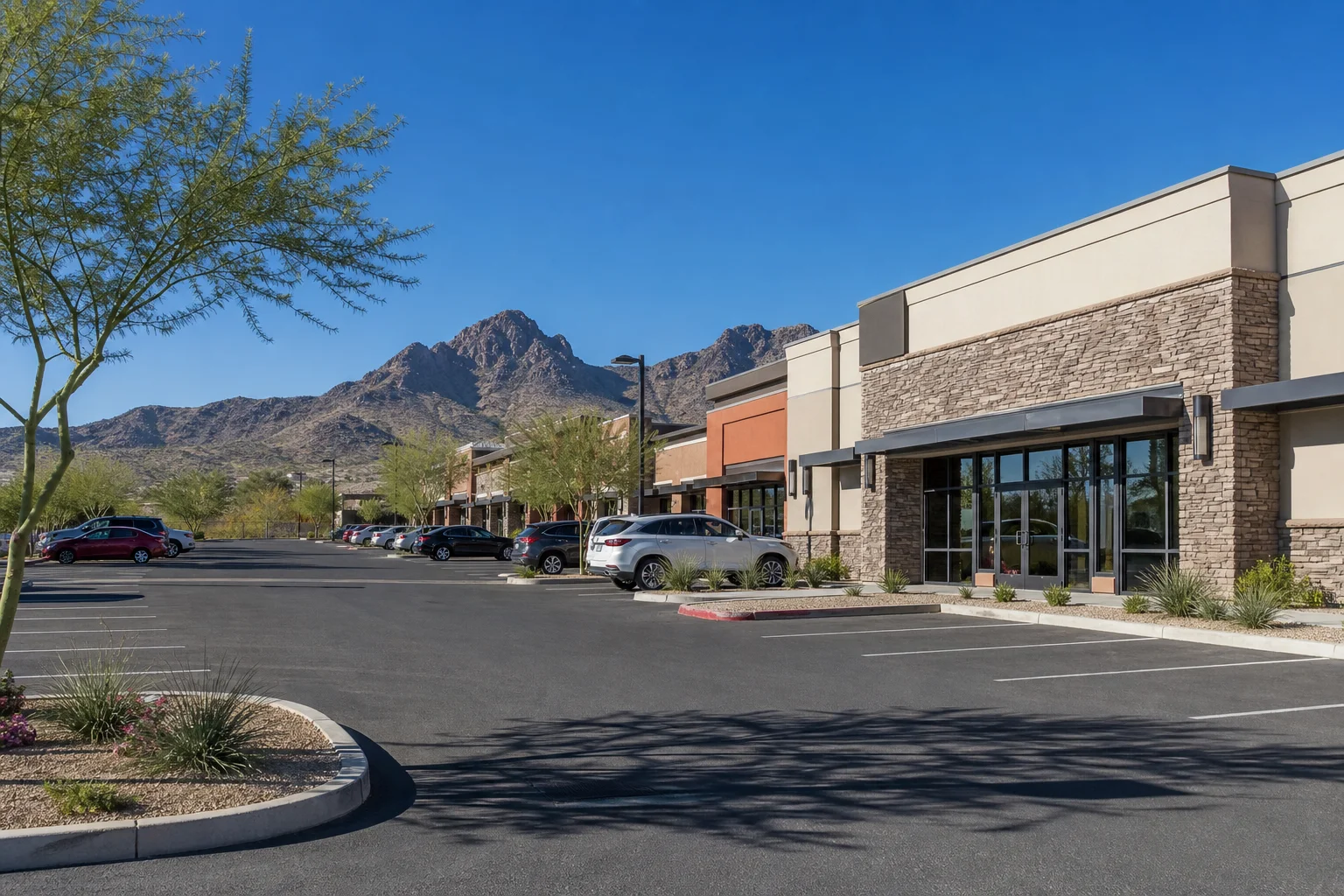 Aerial view of a Phoenix metro anchored retail shopping center with full parking lot, modern storefronts, and Arizona desert skyline in the background, photographed in clear daylight
